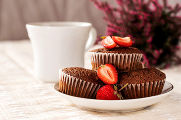 Chocolate muffins with strawberries on a saucer with a white cup of coffee on a white tablecloth with pink heather in the background.