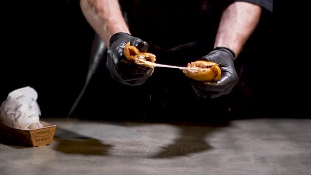 Professional chef cooker in black gloves breaks into two pieces tasty hot fried grilled cheese bun isolated on black background. Slow motion