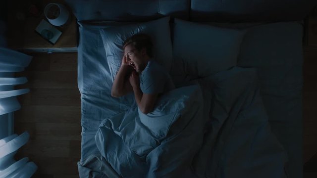 Top View of Handsome Young Man Sleeping Cozily on a Bed in His Bedroom at Nigh, He Tosses and Turns. Blue Nightly Colors with Cold Weak Lamppost Light Shining Through the Window.