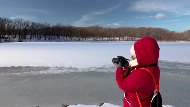 Girl using a DSLR camera taking photos of a landscape near a frozen lake in winter.It's a bright cloudless sunny day with blue sky.