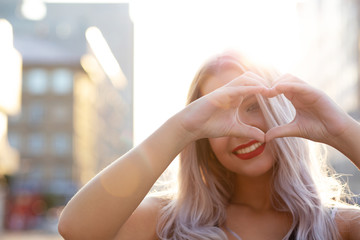 Joyful blonde model with red lips making heart shape with her fingers at the street. Empty space