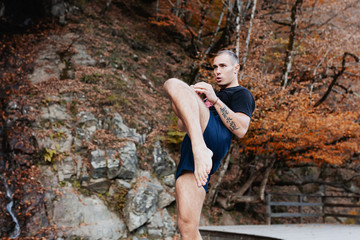 Young strong man in sportswear and barefoot is standing on wooden platform and is practicing kickboxing against background of stone wall and beautiful nature. Health and sport concept. Copyspace