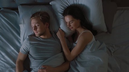 Top View Bed at Night: Attractive young Couple Sleeping Together, Holding Each other in Arms, Embracing. First Rays of Morning Sun Illuminate Room Trough the Window