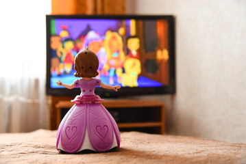 Toy doll in a pink dress is watching a cartoon on the TV