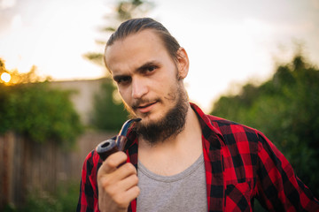 A rustic man with a beard and a red shirt in a cage. A young hipster guy with a pipe in his hand.