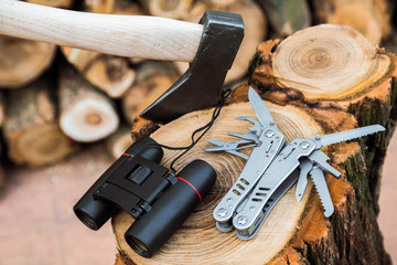 axe, binoculars and multitool knife case on the stump, firewoods on the background