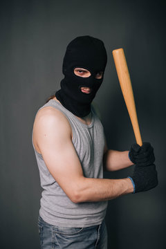 Bandit, the criminal in a black mask and in a gray T-shirt with a painless bat. Robber with a weapon. A man in a balaclava and a gray mask on a gray background