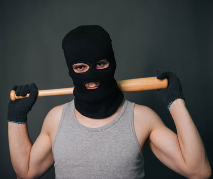 Bandit, the criminal in a black mask and in a gray T-shirt with a painless bat. Robber with a weapon. A man in a balaclava and a gray mask on a gray background
