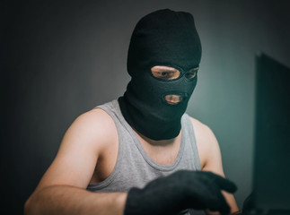 A thief in a black mask stole a laptop. A man in a balaclava and a computer in hand on a gray background. The hacker is hacking the computer. Steals information. A young man breaks the law.