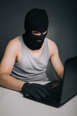 A thief in a black mask stole a laptop. A man in a balaclava and a computer in hand on a gray background. The hacker is hacking the computer. Steals information. A young man breaks the law.