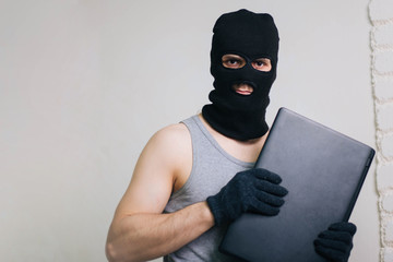 A thief in a black mask stole a laptop. A man in a balaclava and with a computer in hands on a white background. The hacker is hacking the computer. Steals information. A young man breaks the law.
