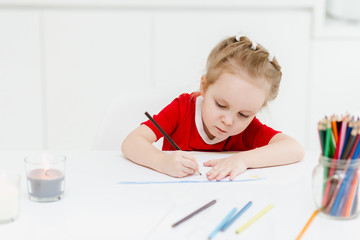 Naklejka premium Cute little Caucasian girl in casual red dress draws with colored pencils in a bright room. Preparing a preschooler for school