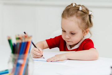 Cute little Caucasian girl in casual red dress draws with colored pencils in a bright room. Preparing a preschooler for school