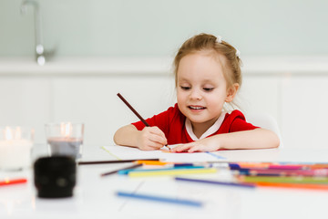 Cute little Caucasian girl in casual red dress draws with colored pencils in a bright room. Preparing a preschooler for school