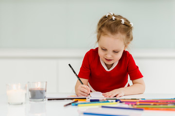 Cute little Caucasian girl in casual red dress draws with colored pencils in a bright room. Preparing a preschooler for school