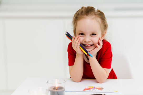 Cute little Caucasian girl in casual red dress draws and  sits at the table and holds colored pencils in his hands. Pozing amd making funny faces. Preparing a preschooler for school