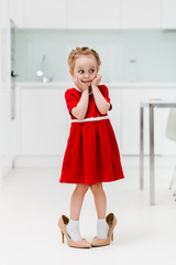 Cute smart little Caucasian fashionable girl in red dress put on her mother's shoes and posing for the camera in a modern minimalist kitchen