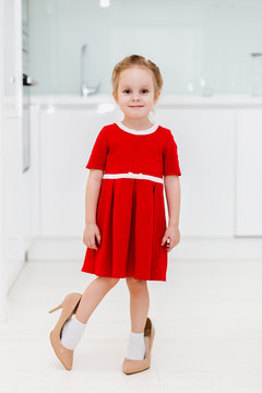 Cute smart little Caucasian fashionable girl in red dress put on her mother's shoes and posing for the camera in a modern minimalist kitchen