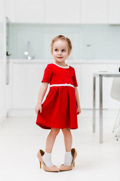 Cute smart little Caucasian fashionable girl in red dress put on her mother's shoes and posing for the camera in a modern minimalist kitchen