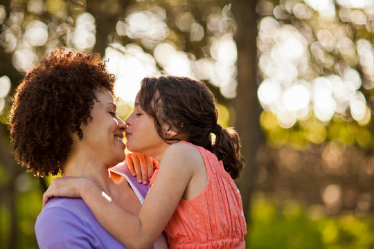 Smiling mid adult woman being kissed by her young daughter as she holds her in the forest.