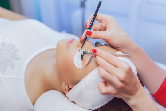 Eyelash Extension Procedure. Close up view of beautiful Woman with Long Eyelashes. Stylist holding tweezers, tongs and making lengthening lashes for girl in a beauty salon. Beauty Concept.