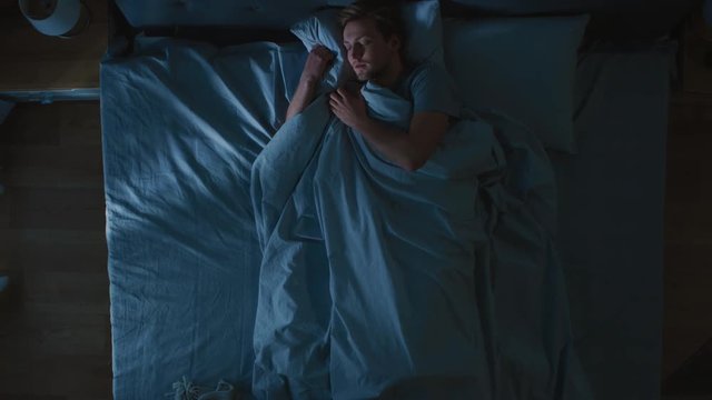 Top View of Handsome Young Man Sleeping Cozily on a Bed in His Bedroom at Night. Blue Nightly Colors with Cold Weak Lamppost Light Shining Through the Window.
