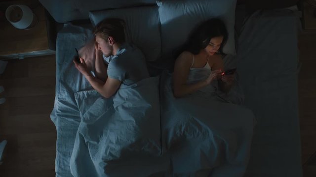 Alienated Millennial Young Couple in the Bed, Young People Turn Away From Each other Using Smartphones, Browsing Through Social Networks and Not Talking to Each Other. Top Down Camera Shot