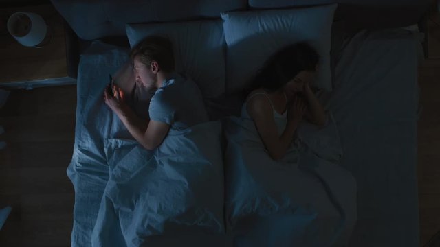 Insomniac Stressed Young Man Uses Smarphone at Night in Bed when Girlfriend Sleeps Sweetly and Soundly. Top View Camera