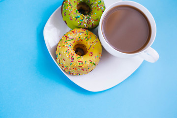 Two colored donuts on a plate with a cup of coffee. Yellow and Green Donut on a blue background. Dessert