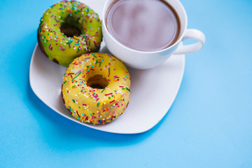 Two colored donuts on a plate with a cup of coffee. Yellow and Green Donut on a blue background. Dessert