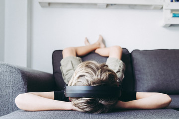 Blond boy barefoot legs crossed listening to music with headphones, spend free time relaxing singing Student lying on its back comfortably relaxed at home. Domestic scene bottom view Arms behind head