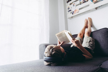 Blonde boy barefoot reading book student lying down learn the lesson comfortably relaxed diligent child reading a story young man plunges into culture Read and listen to music with headphones at home