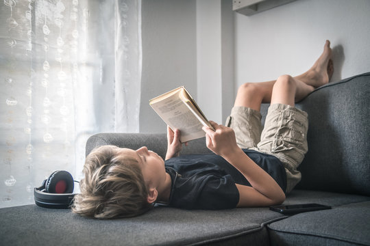 Blonde boy barefoot reading book student lying down learn the lesson comfortably relaxed diligent child reading a story young man plunges into culture Read and listen to music with headphones at home