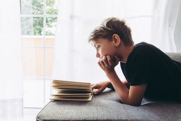 Blond boy reads book with his face leaning on a hand, student lying down reads the lesson comfortably relaxed, diligent child reads a story, young man plunges into culture Home works on sofà backlight