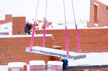 Reinforced concrete slab, crane transportation. Tape slings are used. Place of action - construction of a brick house. The visible construction workers in hard hats. Winter, lies snow.