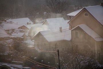 Smoking chimneys at roofs with snow of houses emits smoke, smog at sunrise, pollutants enter atmosphere. Environmental disaster. Harmful emissions, exhaust gases into air. Winter day, heating season.