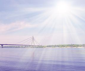 The bright sun shines with glare over the river and the bridge in the city.