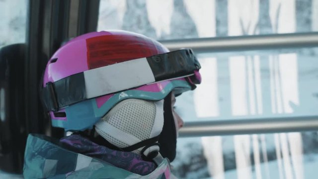 Medium close-up shot of a cute young girl in full skiing gear looking out the window of a ski lift.