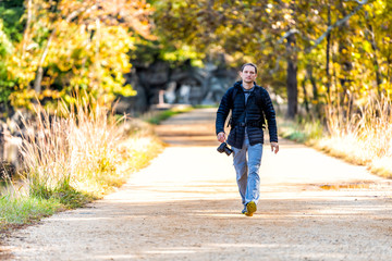 Young photographer man walking on trail path road by trees during autumn Potomac river canal in Great Falls, Maryland with colorful foliage sunny sunlight