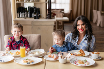 Beautiful young woman with her baby sons. Light breakfast near window in a cafe. Croissants, omelet, coffee and many different dishes on the table in the cafe. Happy family