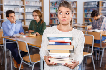 Annoyed girl sitting with pile of books in high school library on background of working fellow students