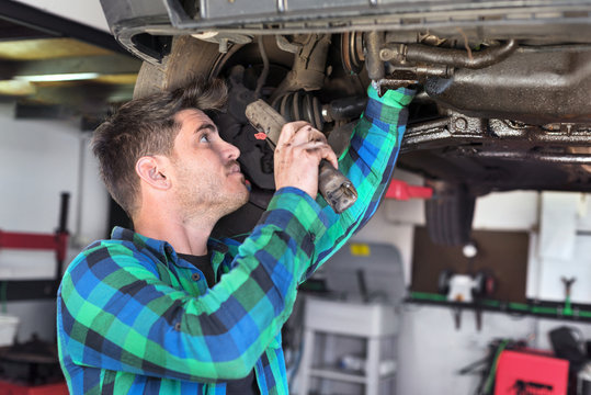 Handsome car mechanic checking suspension system of a lifted car at repair service station .
