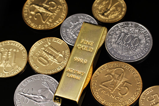 A macro image of an assortment of West African Franc coins and a gold one ounce ingot on a reflective black background