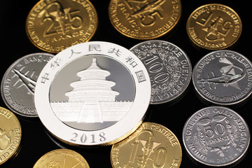 A macro image of an assortment of West African Franc coins and a silver Chinese one ounce coin on a reflective black background