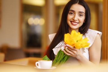 Obraz na płótnie Canvas Beautiful Young Woman With Spring Tulips Flowers Bouquet At Cafe. Woman Drinking Coffee And Using Phone. Spring Style. Brunette Woman In Nice Spring.Beautiful Spring Mood. Fashion Spring/Summer.