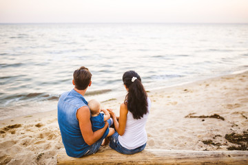 Theme family weekend, beach summer vacation with child. Young stylish family of three dad mom and daughter sit one year with their backs on lying tree, log overlooking horizon sea sunset in summer