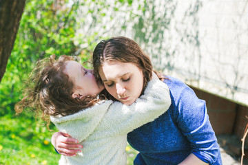 Fototapeta premium Little girl hugs mother sitting on the grass. Child, playing, threw his mother on grass. Family in spring walks on street.