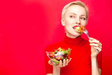 Beautiful woman eating salad wearing red dress and red lisptick over red background. Holiday, ...