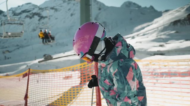 A young girl adjusts her gear at the top of a ski hill. Ski lifts go by in the background.