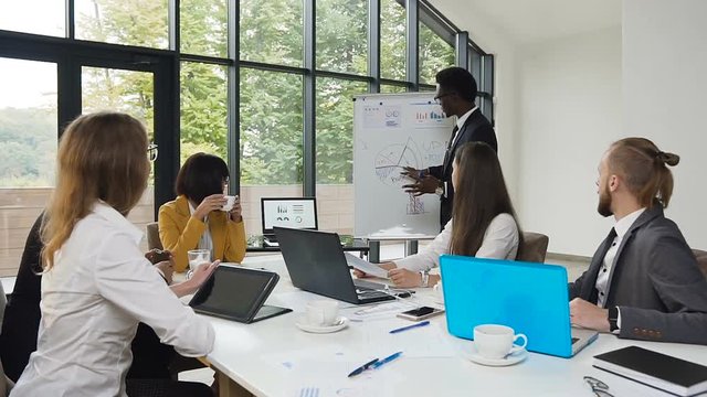 African american man drawing diagram on flipchart and holding to presentation for business colleagues at the corporate training in office with panoramic windows to the park